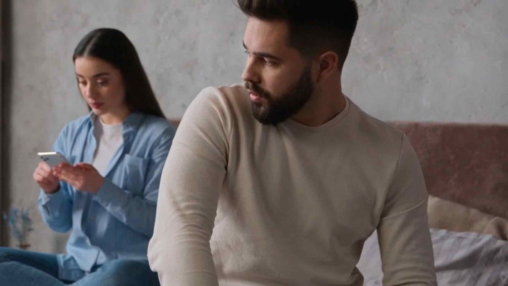 Suspicious man turning to look at a woman sitting behind him on a bed, who is focused on her phone